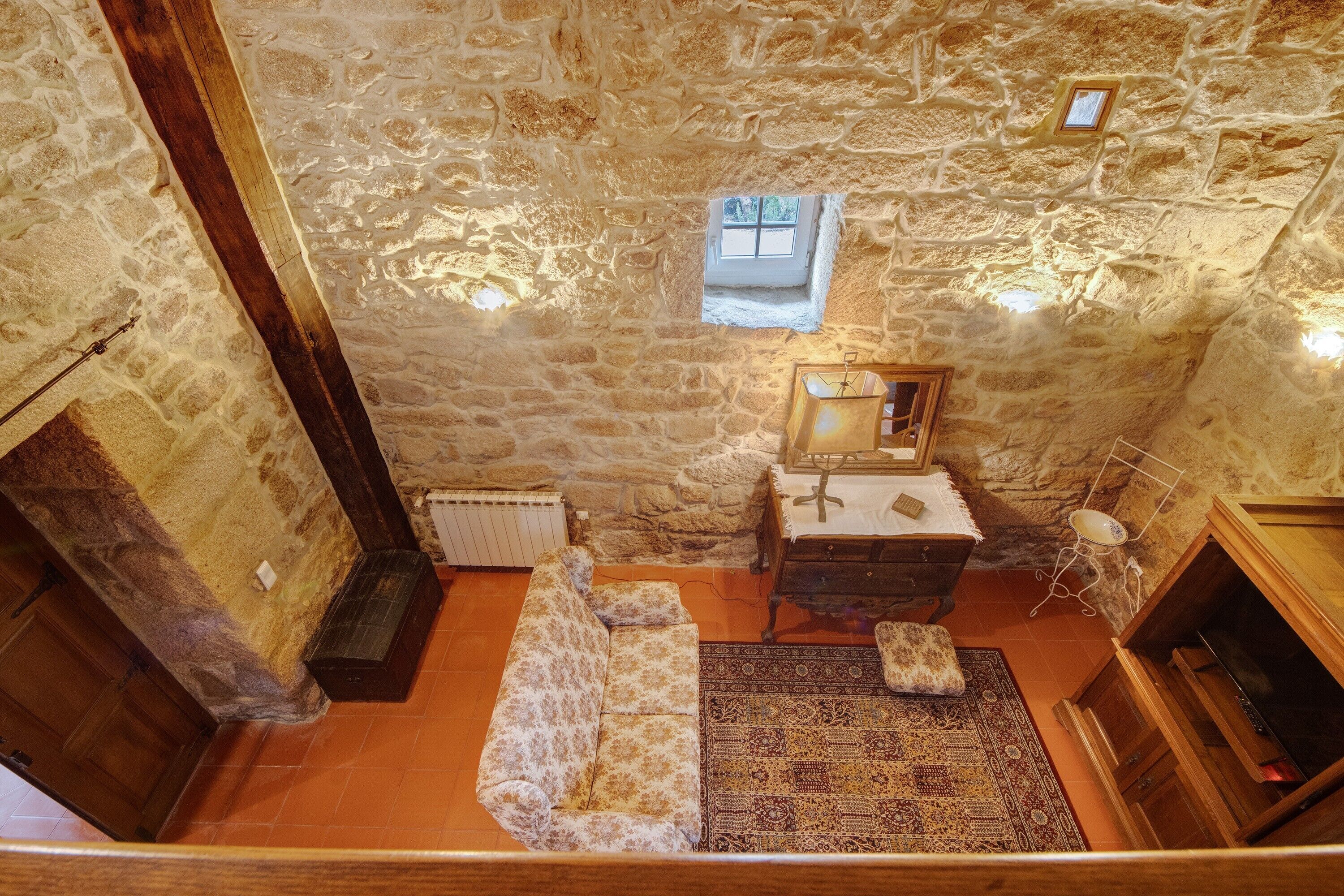 Une pièce aux murs de pierre, avec un banc en bois, une petite table surmontée d’un miroir et un tapis à motifs.