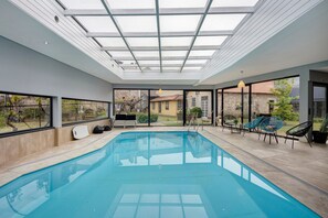 Piscina interior moderna con techo de vidrio, ventanas amplias y zona de estar exterior.
