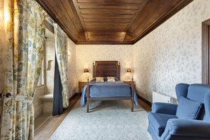 Un dormitorio con techo de madera, una cama con cabecera, dos mesitas de noche con lámparas, un sillón azul y cortinas estampadas.