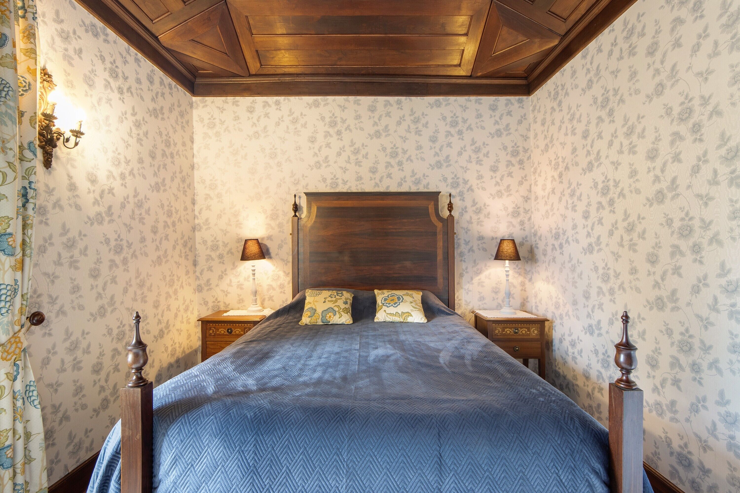 A bedroom with a wooden headboard, two bedside lamps, floral wallpaper, and a patterned bedspread.