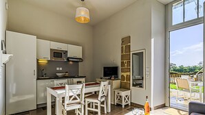 A modern kitchen with white cabinets, a dining area with white chairs, a table with a bottle and snacks, and a large window with a view of the outdoors. - Le Château de Kergonano by Interhome (Baden)
