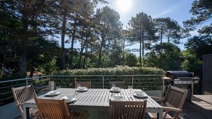 An outdoor dining area with a wooden table and chairs, surrounded by a deck with a view of tall trees and a grill. - Holiday home Le Penon – Terrace, balcony, Wi-Fi, 6 guests, close to Seignosse beach (Seignosse)