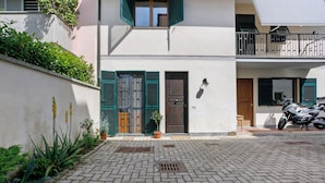 Une cour pavée avec un pot de fleurs, une moto et un bâtiment doté d’une porte et de volets.