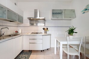 Cocina compacta con estufa a gas, campana extractora de acero inoxidable, muebles blancos y una zona de comedor con mesa y sillas.
