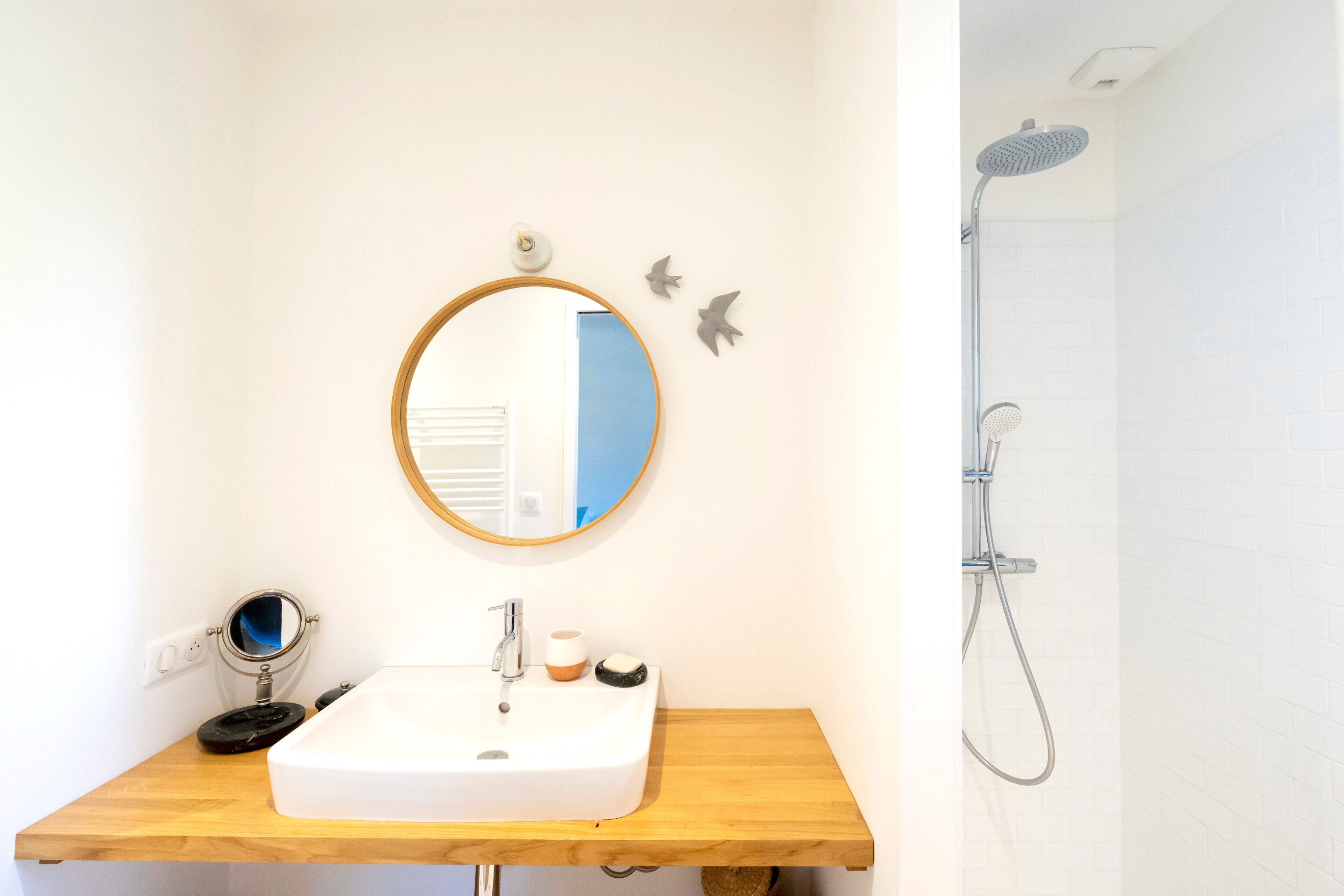 Une salle de bain avec un meuble-lavabo en bois, un miroir rond, un évier, un miroir à main, un porte-brosse à dents, une brosse à dents, un gobelet, un porte-savon et une douche avec un pommeau de douche à main.