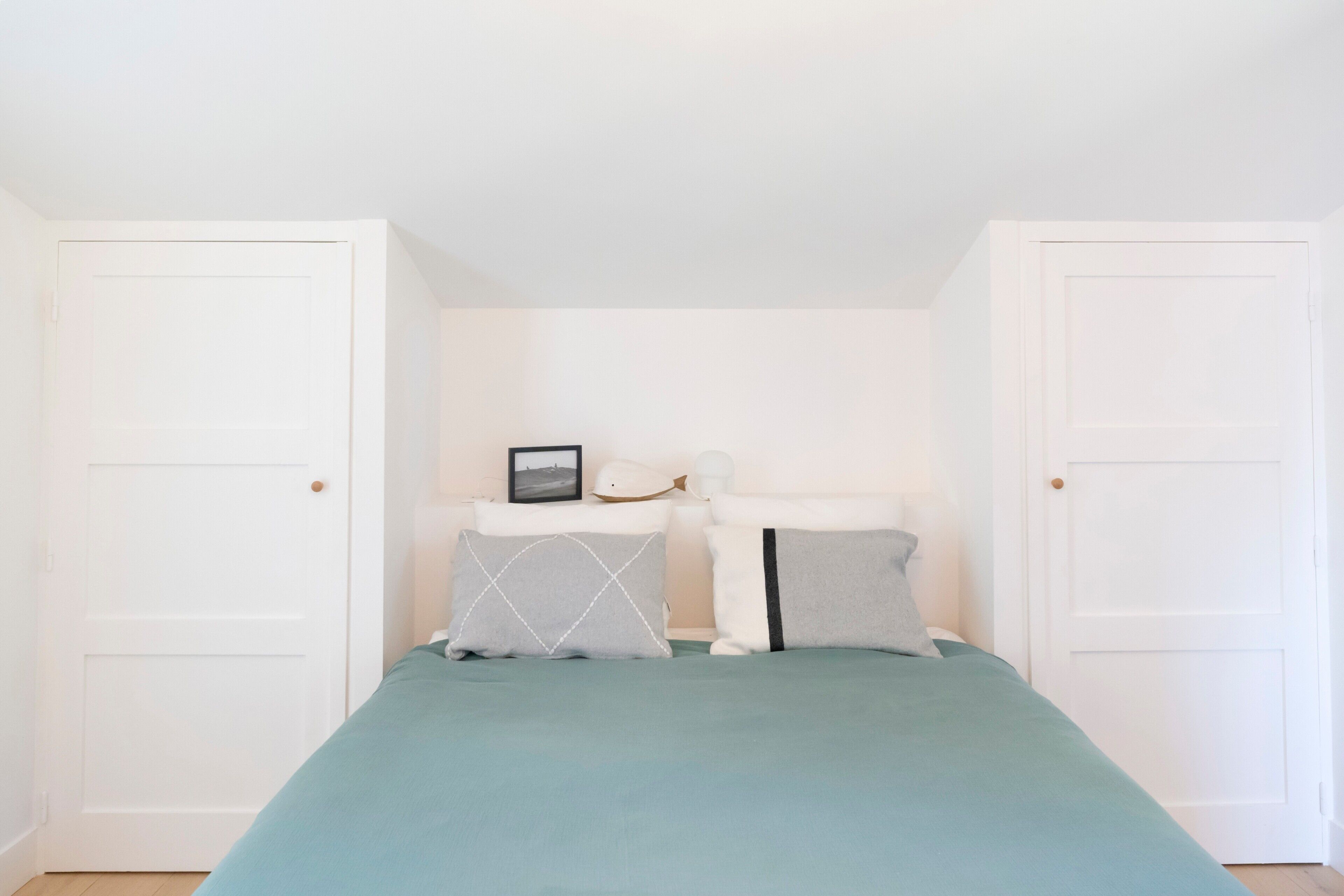 A bedroom with a bed, two white doors, a nightstand with a picture frame, and a lamp.