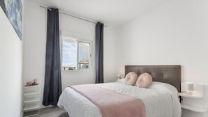 Un dormitorio con cama, cortinas, mesita de noche, estante en la pared y ventana.