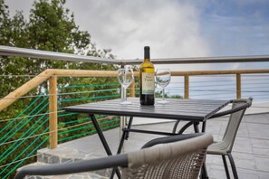 Uma mesa com uma garrafa de vinho e duas taças numa varanda externa com vista para o mar.