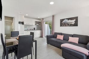 Uma sala de estar moderna com um sofá cinzento, uma mesa de jantar com cadeiras, uma área de cozinha e uma janela grande com cortinas.