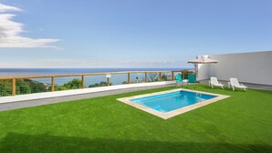 Una piscina en la azotea con vista al océano, rodeada de césped artificial y sillones blancos para tomar el sol.
