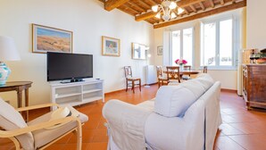 Un salon comprenant un téléviseur, un canapé blanc, des chaises en bois, une table à manger avec des chaises et un lustre.