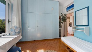 A room with a bed, a blue wardrobe, a fan, a table, and a view of the outdoors.