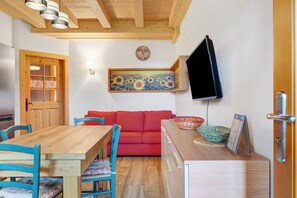 Une pièce comprenant une table à manger en bois, des chaises bleues, un canapé rouge, un téléviseur fixé au mur et un meuble en bois sur lequel est posé un calendrier.