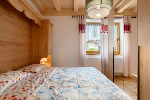 A bedroom with wooden walls, a bed with floral and blue patterned bedding, a bedside lamp, and a window with curtains. - Apartment 'Piano Terra 3' with Mountain View, Private Terrace and Wi-Fi (Romeno)