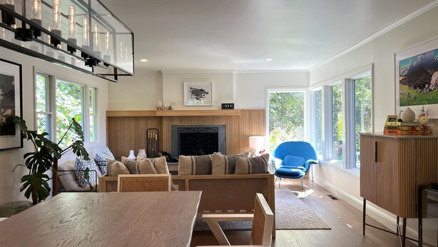 A modern living room with a wooden dining table, a sofa, and a blue chair. There is a fireplace, a large window, and a plant. - Storybook Summer Stay - 4-bedroom house in charming Mill Valley (Mill Valley)
