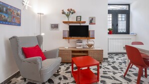 Ein Wohnzimmer mit einem Hundszahn-Sessel, einem roten Tisch, einem Flachbildfernseher und einem an der Wand befestigten Regal mit Blumen.