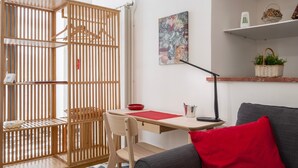 Ein Zimmer mit Schreibtisch, Stuhl und einem grauen Sofa mit rotem Kissen. Es gibt eine Holzwand mit Kleiderstange, ein Bild an der Wand und ein Regal mit Dekoration.