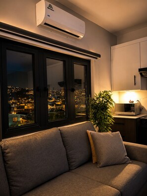 Uma sala de estar moderna com um sofá, uma planta em vaso e vista para o horizonte urbano através de grandes janelas.