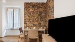 Una habitación con pared de piedra, una mesa con sillas, un televisor y una ventana con cortinas.