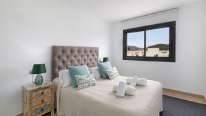 Ein Schlafzimmer mit einer gebügelten Kopfteil, einem Bett mit weißen Leinen, Kissen in Türkis und Weiß mit Muster, einem kleinen Nachttisch mit einer Lampe und Blick auf Gebäude durch ein Fenster.