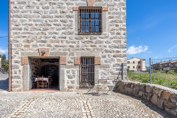 Un bâtiment en pierre avec une cour pavée, une table et des chaises en bois à l’extérieur, et une fenêtre avec des barreaux en métal.