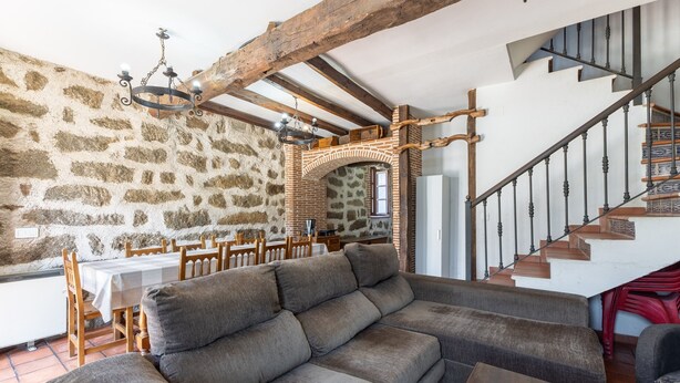 Una habitación con pared de piedra, escalera de madera, sofá y mesa de comedor.
