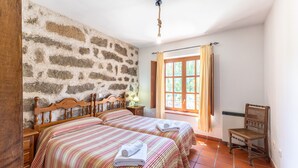 Una habitación con dos camas, una silla de madera, una pared de piedra, una ventana con cortinas y una lámpara en el techo.
