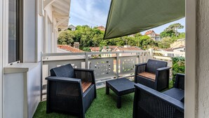 Un balcon doté de meubles en osier, d’un sol recouvert d’un gazon synthétique vert et offrant une vue sur les maisons au loin.