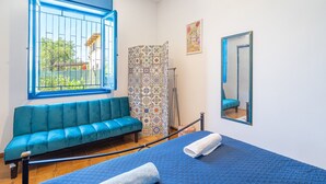 Una habitación con una cama, un sofá azul, una ventana con marco azul, un rincón de ducha y un espejo.