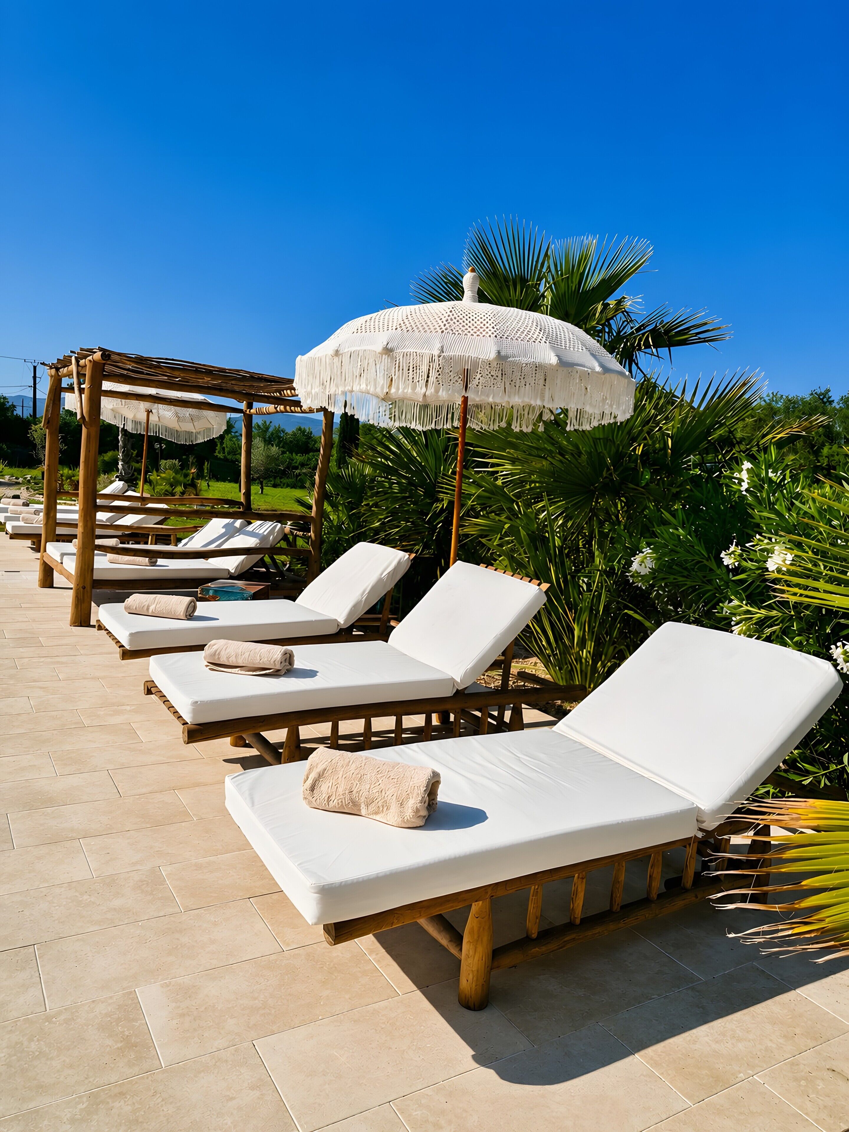Une rangée de chaises longues avec des coussins et des serviettes blancs, une pergola en bois et un parasol blanc à franges.