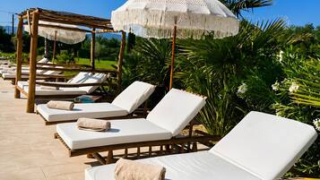 Une rangée de chaises longues avec des coussins et des serviettes blancs, une pergola en bois et un parasol blanc à franges.