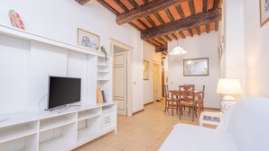 Una habitación con una televisión sobre un estante blanco, un área de comedor con sillas y una mesa, y una cama con cabecera blanca.