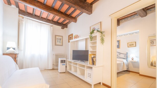 Un dormitorio con una cama, un televisor sobre un soporte blanco, un radiador y una ventana con cortinas.