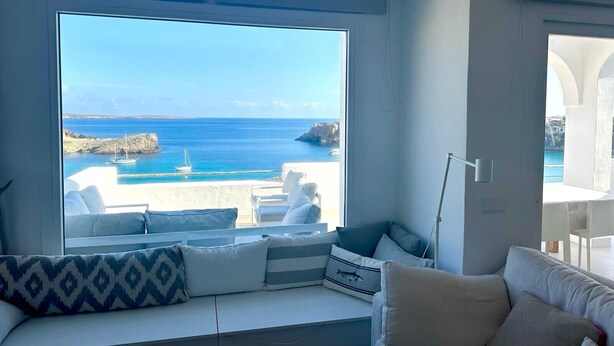 Habitación con un ventanal grande que ofrece vistas al mar, un sofá blanco con cojines y una pared blanca.