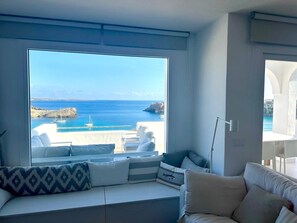 Ein Zimmer mit großem Fenster und Blick auf die Küste, ein weißes Sofa mit Kissen und eine weiße Wand.
