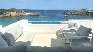 Terraza con muebles blancos al aire libre que da a una zona costera con aguas azules cristalinas y terreno rocoso.
