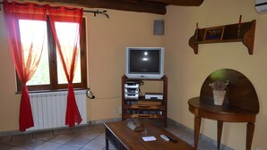 Ein Wohnzimmer mit einem Fernseher auf einem Schrank, einem Holztisch, einem kleinen Tisch mit einer Vase und einem Fenster mit Vorhängen.