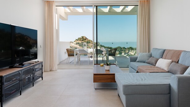 A modern living room with a flat-screen TV, a sofa, a coffee table, and a balcony with a view of the sea.