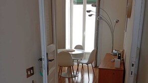 Ein schmales Entree führt zu einem Essbereich mit Tisch und Stühlen, einem Schreibtisch mit Lampe sowie einem Fenster mit Jalousien.