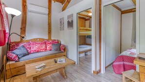 Ein gemütliches Zimmer mit Holzbalkendecke, einer Couch mit gemusterten Kissen, einem kleinen Tisch mit Fernbedienung und Zeitschrift sowie einem Etagenbett mit roter und weißer Steppdecke.