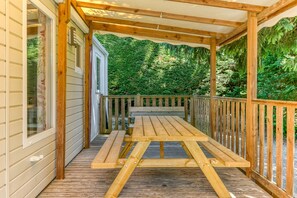 A wooden deck with a picnic table and benches, surrounded by a wooden railing, with a house and dense greenery in the background.