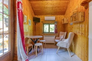 Interior de una cabaña de madera con una mesa redonda, dos sillas, un sofá y una ventana con cortinas.