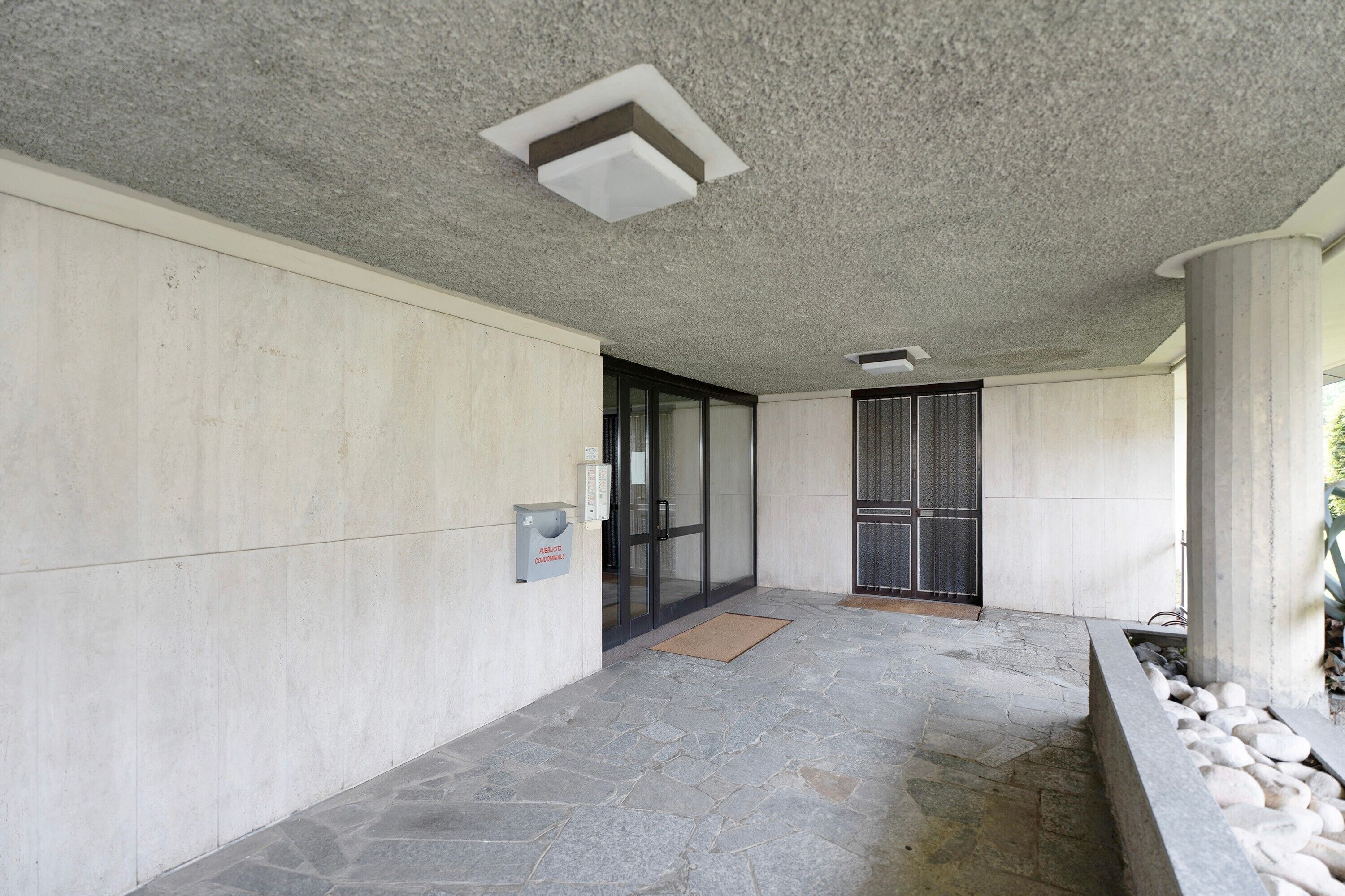 A covered outdoor area with stone flooring, a concrete wall, a glass door entrance, and a small planter with rocks.