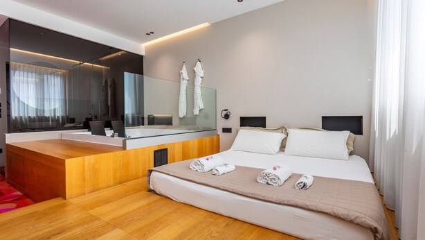 Une chambre d’hôtel moderne avec un bureau en bois, un grand miroir, un lit avec des serviettes soigneusement pliées et une grande fenêtre avec des rideaux.
