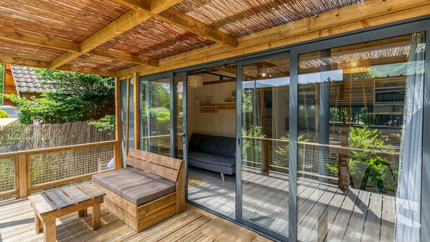 A wooden deck with a bench and a sofa, a glass door leading to a balcony, and a view of greenery.
