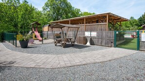 Een overdekte picknickplaats met een houten bankje, een speeltuin met een glijbaan en een omheinde tuin.