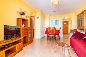 Uma sala de estar com televisão, móveis de madeira, uma área de jantar com cadeiras e uma mesa, e um sofá vermelho.
