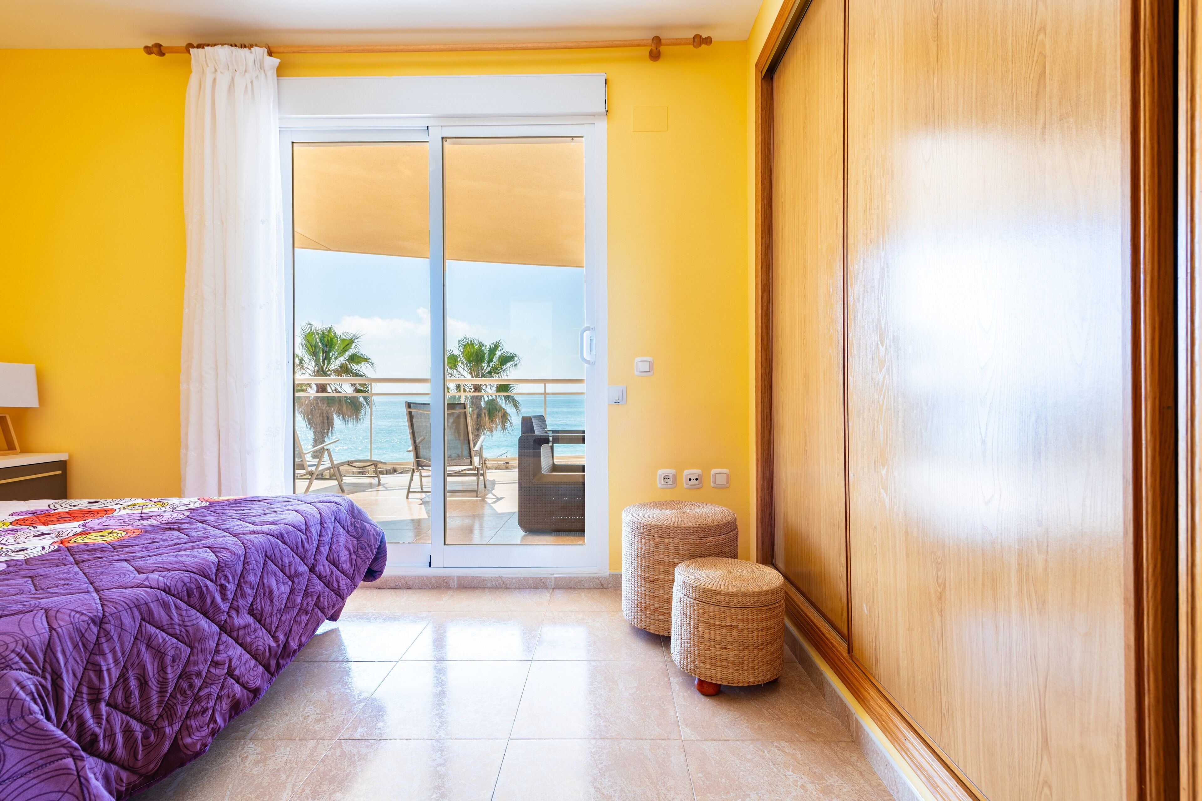 Une chambre d’hôtel avec un lit, une porte-fenêtre coulissante donnant sur un balcon et deux poufs en osier.