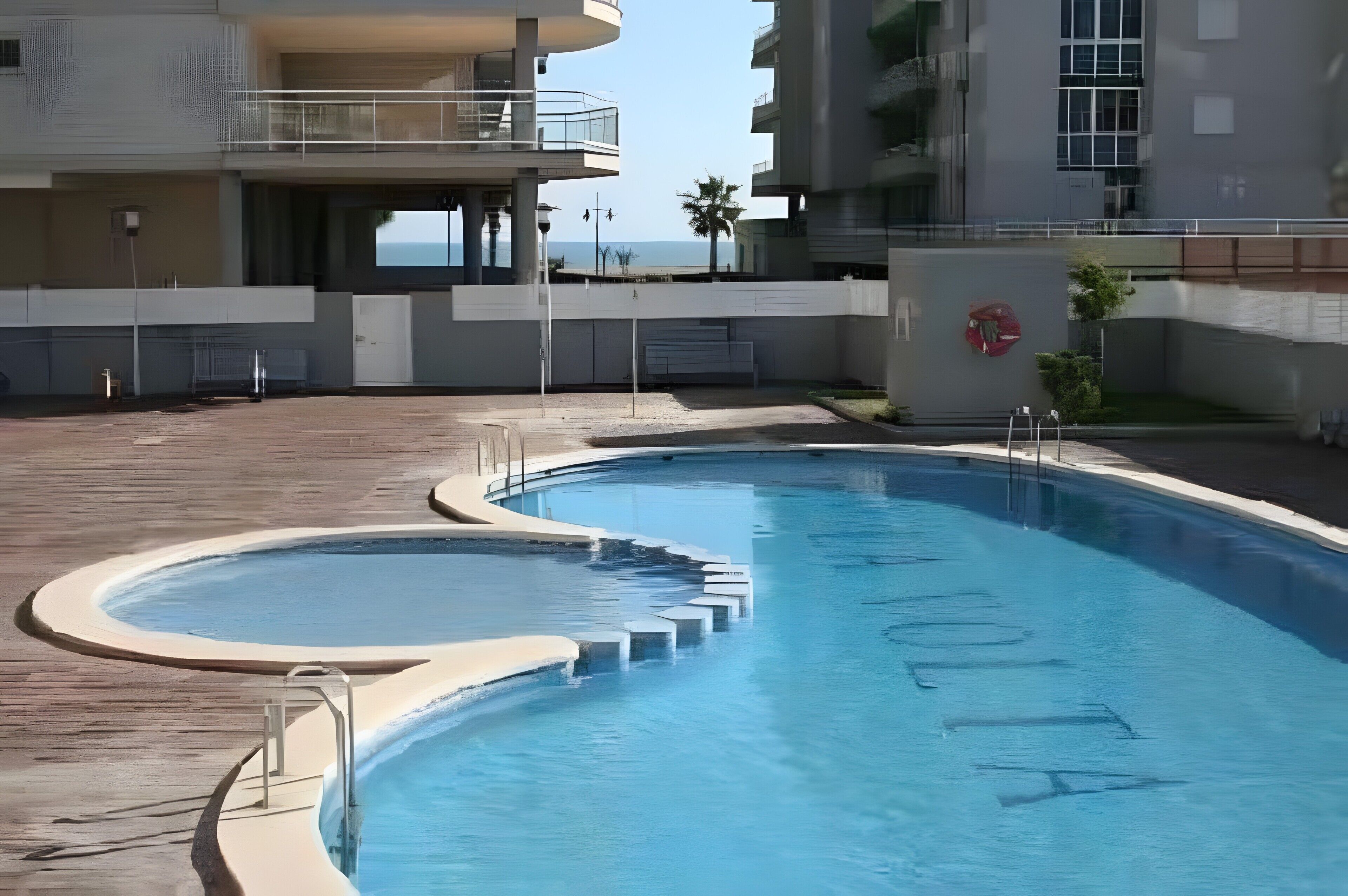 A swimming pool with a curved design, surrounded by a wooden deck and a building with balconies.