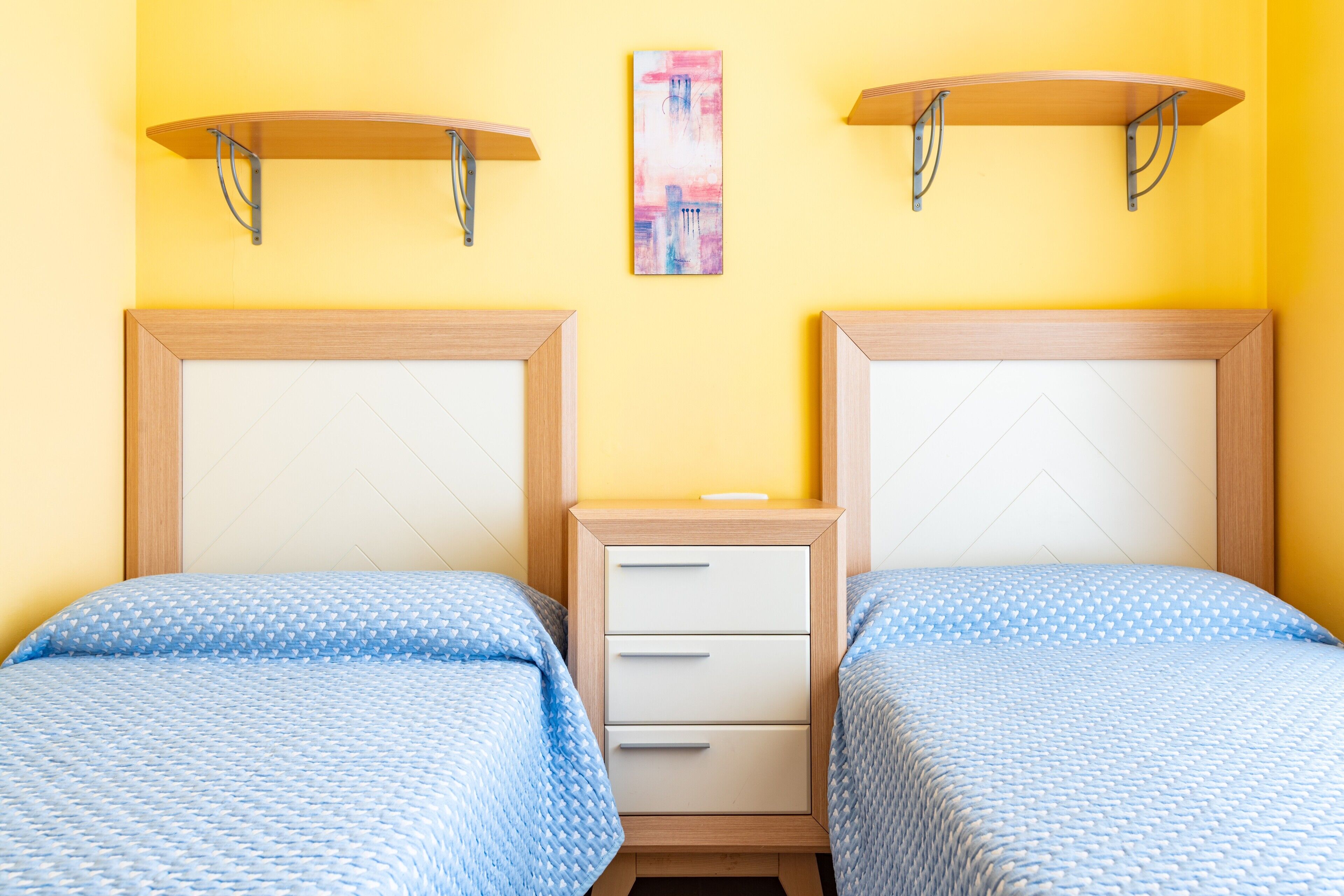 A room with two beds, a nightstand, and a wall-mounted shelf.